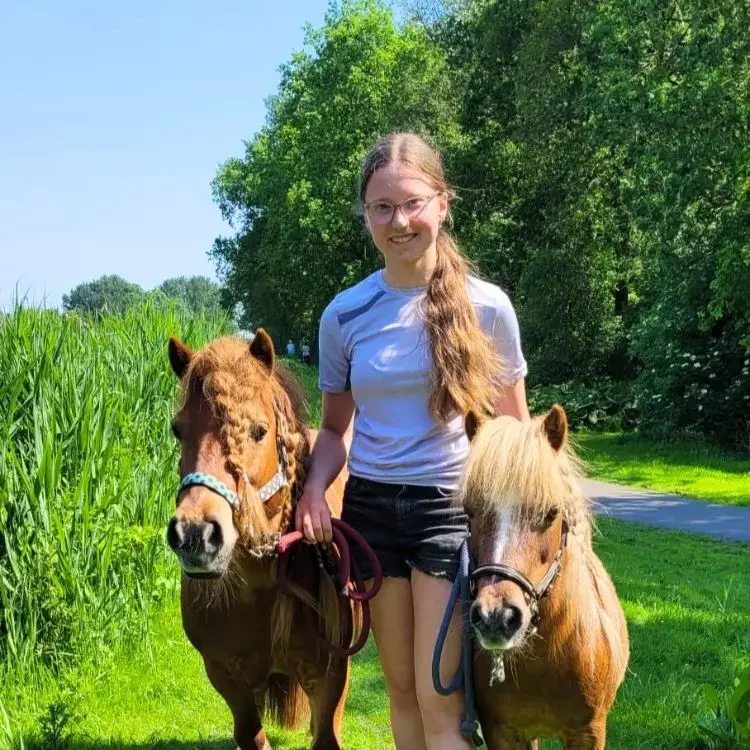 Eline staat met twee pony’s in een groene buitenomgeving, glimlachend en verbonden met de dieren op een zonnige dag
